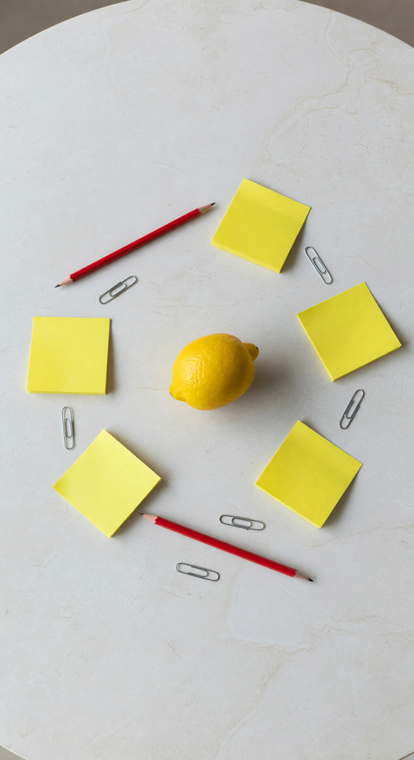 Grok Imagine Image output of a top-down flat lay with red pencils, paper clips, a lemon, and yellow sticky notes arranged in a spiral